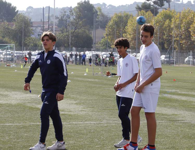 Trois jeunes joueurs de football sur un terrain d'entraînement, avec d'autres personnes en arrière-plan.