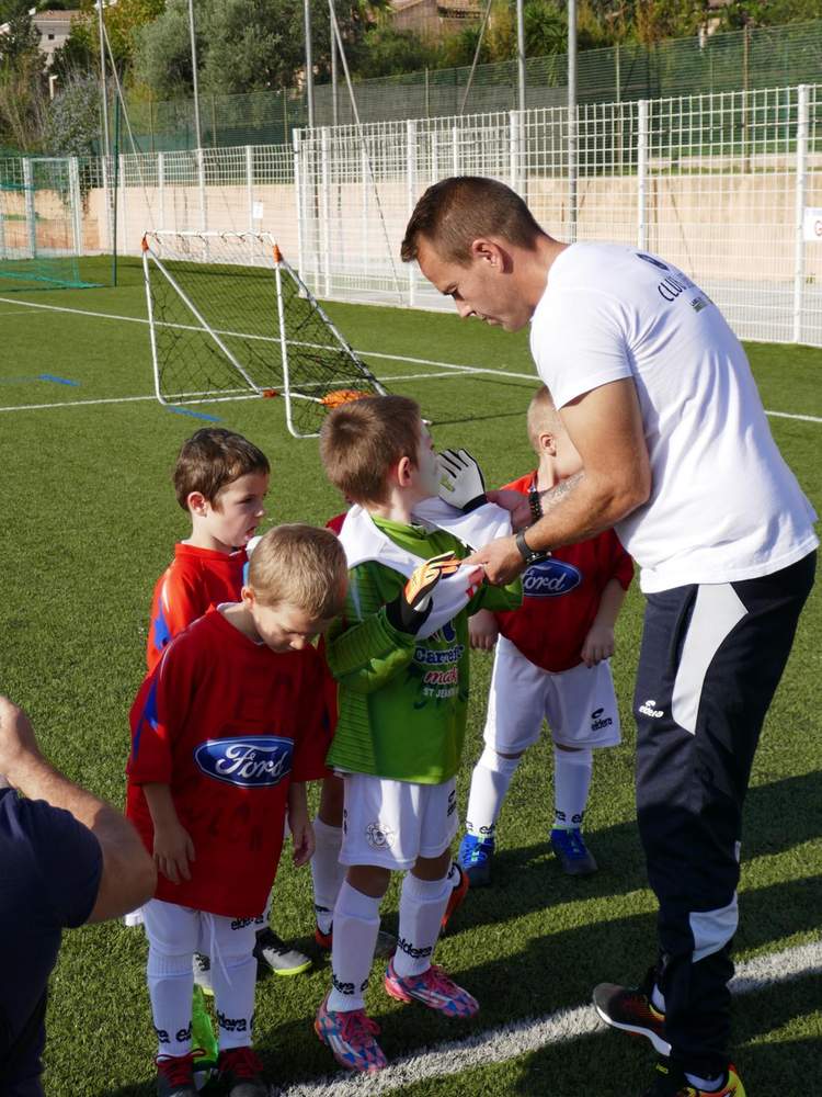 Un entraîneur aide un jeune joueur à enfiler ses gants de gardien sur un terrain de football synthétique.