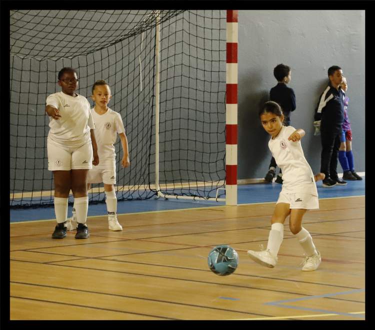 Des jeunes joueurs de football en action sur un terrain de futsal, avec une fille en blanc frappant le ballon.