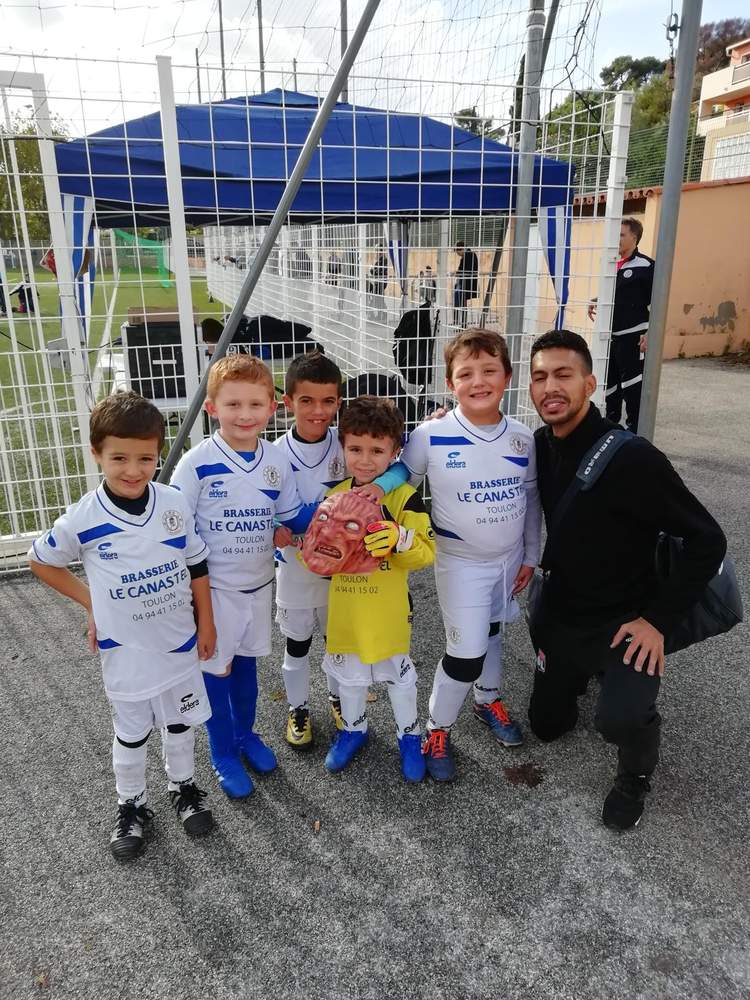Un groupe de jeunes footballeurs en maillot blanc et bleu pose avec un adulte sur un terrain extérieur.
