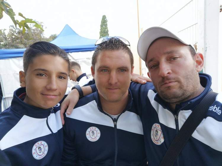 Trois hommes souriants posent ensemble, vêtus de survêtements de club, avec une tente bleue en arrière-plan.