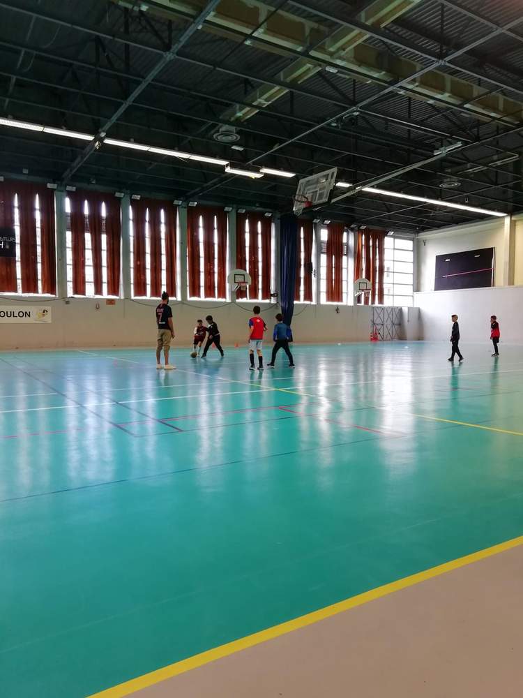 Des enfants jouent au football en salle sous la supervision d'un adulte.