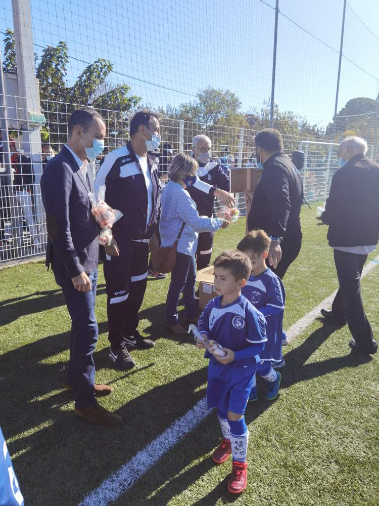 Des adultes masqués distribuent des cadeaux à de jeunes footballeurs sur un terrain ensoleillé.