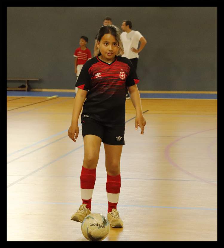 Une jeune joueuse de football en tenue de sport, avec un ballon à ses pieds, regarde l'objectif dans un gymnase.