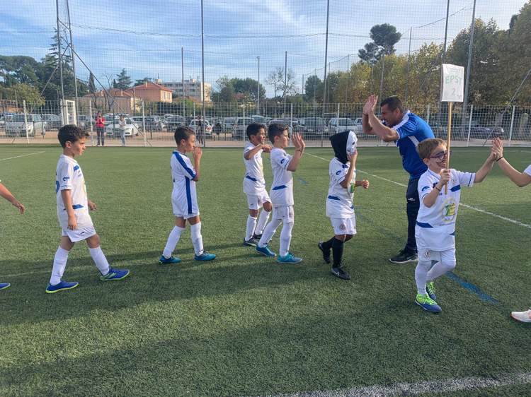 De jeunes footballeurs en tenue blanche et bleue saluent leur entraîneur sur un terrain de football synthétique.