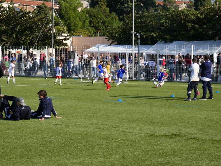 Des jeunes joueurs de football en pleine action sur un terrain en gazon synthétique, avec des spectateurs en arrière-plan.