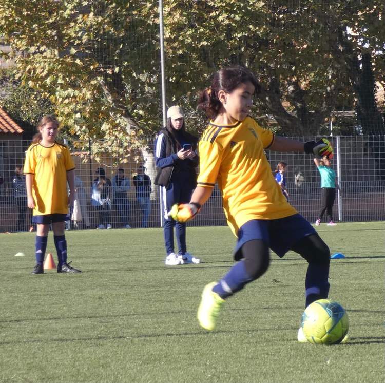 Une jeune joueuse de football en maillot jaune frappe le ballon sur un terrain synthétique ensoleillé.