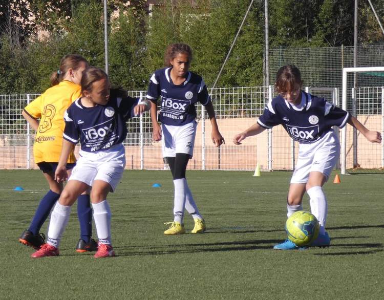 Des jeunes joueuses de football en action sur un terrain vert, avec un ballon au premier plan.