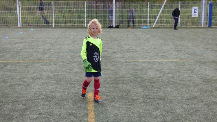 Un jeune gardien de but aux cheveux blonds sourit sur un terrain de football synthétique.