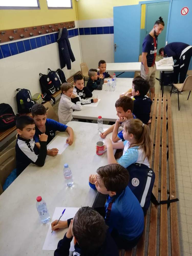 Des jeunes joueurs de football sont assis à des tables, écrivant ou écoutant, sous la surveillance d'adultes.