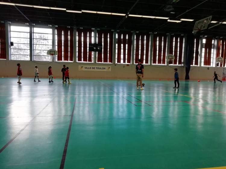 Des enfants s'entraînent au football en salle sous la supervision d'un adulte.