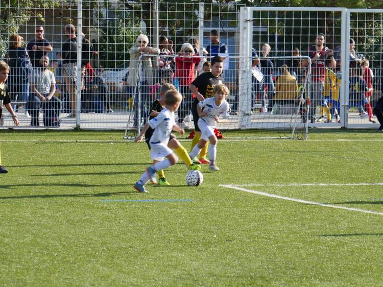 Des jeunes joueurs de football en pleine action sur un terrain ensoleillé, avec des spectateurs en arrière-plan.
