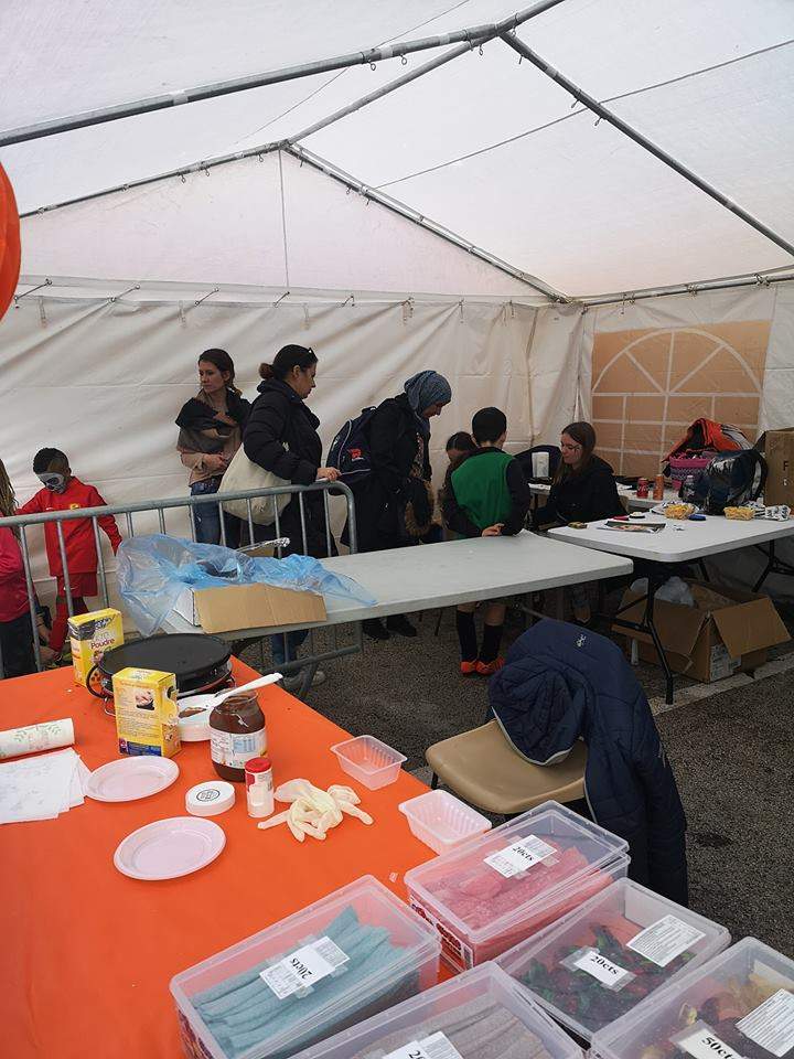 Des personnes se tiennent sous une tente, avec des tables remplies de nourriture et de fournitures au premier plan.