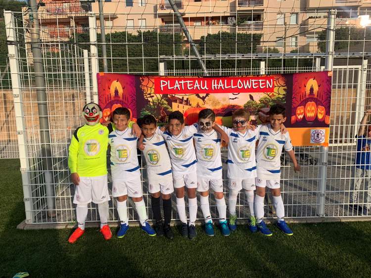 Une équipe de jeunes footballeurs pose devant une banderole d'événement, certains avec des maquillages ou masques d'Halloween.