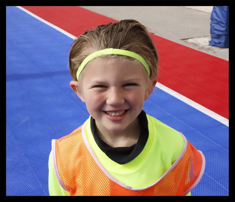 Un jeune joueur souriant porte un maillot fluo et un dossard orange sur un terrain de sport.
