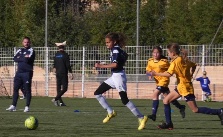 Des jeunes joueuses de football en pleine action sur le terrain, avec des adultes en arrière-plan.