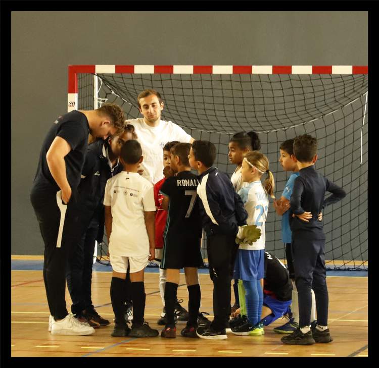 Des enfants et des adultes sont regroupés en cercle sur un terrain de sport en intérieur, devant un but de football.