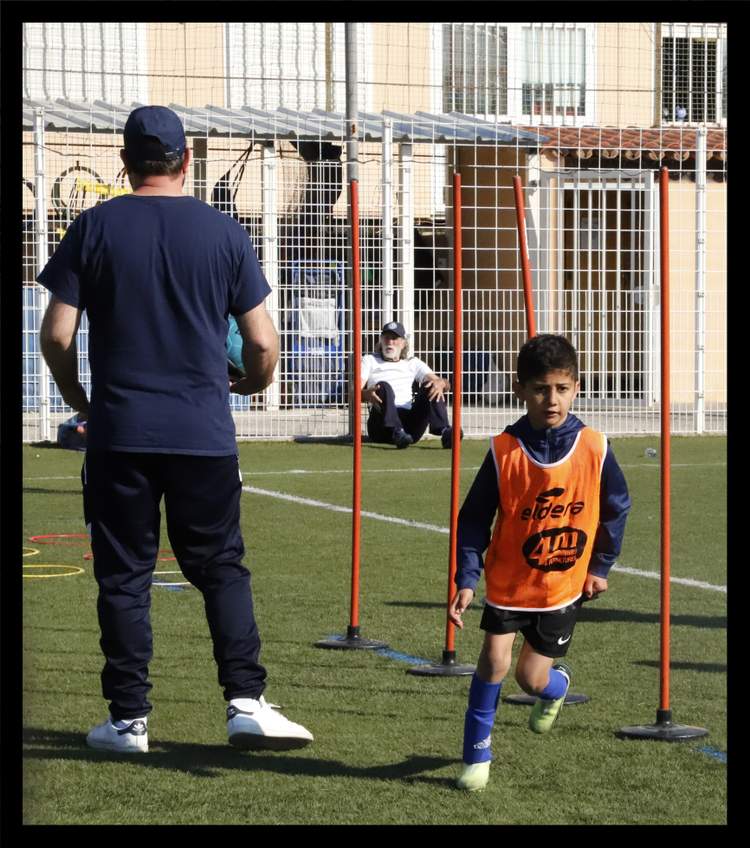 Un jeune joueur en chasuble orange court sur un terrain de football, avec un adulte de dos et un autre assis en arrière-plan.