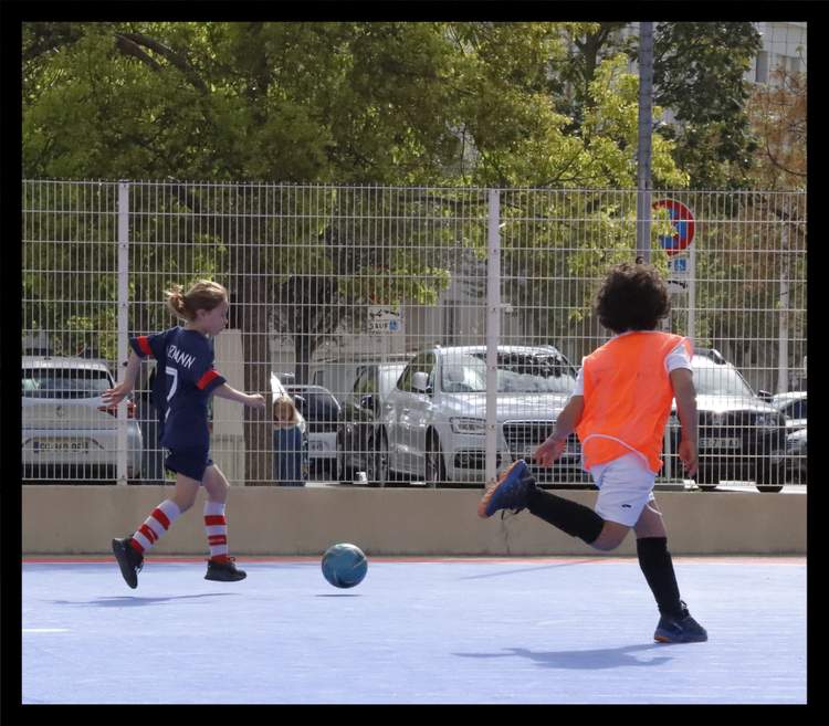 Deux jeunes joueurs de football en pleine action sur un terrain bleu, avec un grillage et des voitures en arrière-plan.