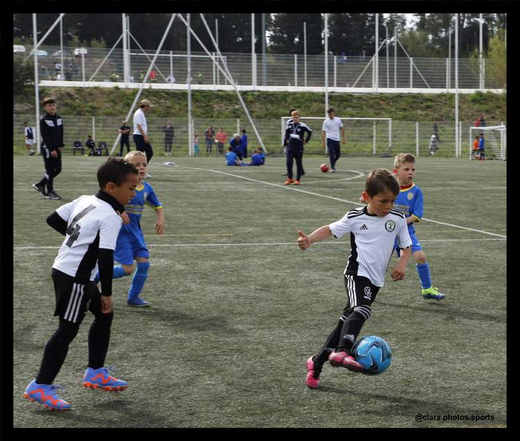 Des jeunes joueurs de football en pleine action sur un terrain en herbe, avec d'autres personnes en arrière-plan.