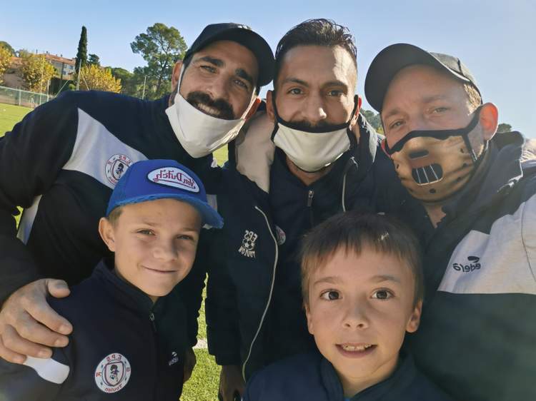 Cinq personnes, dont deux enfants, posent souriantes pour un selfie sur un terrain de football ensoleillé.