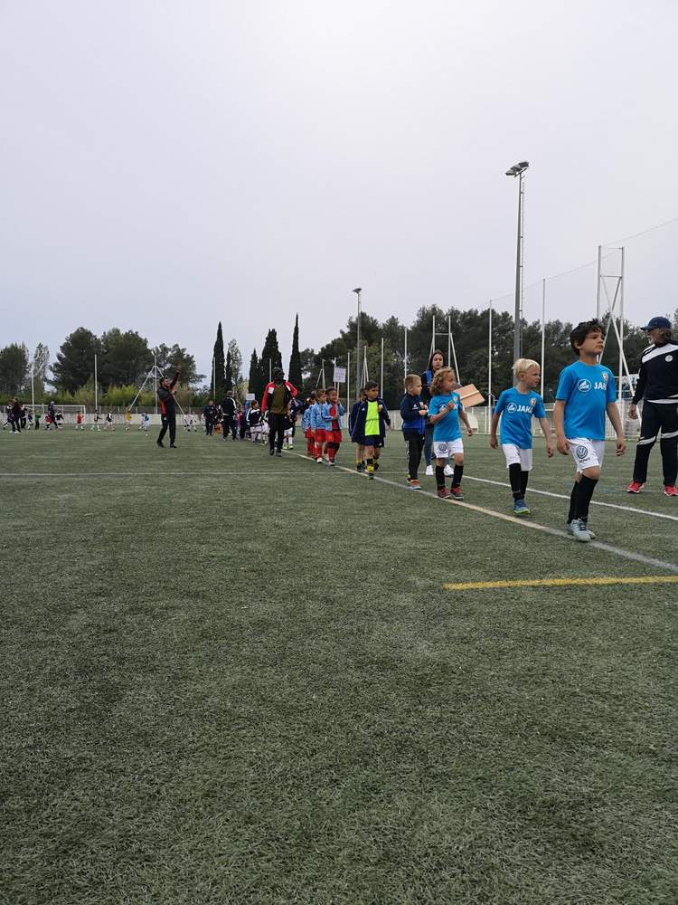 Des jeunes joueurs de football en tenue de sport marchent en ligne sur un terrain synthétique, accompagnés d'adultes.