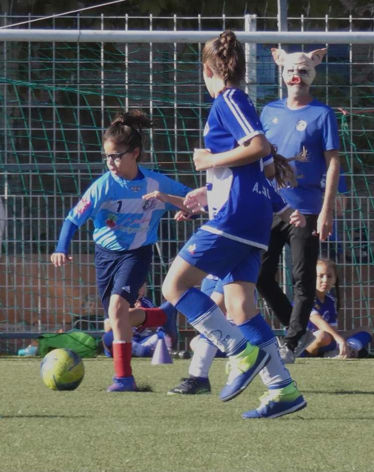 Des jeunes joueuses de football en action sur le terrain, avec un adulte portant un masque de cochon en arrière-plan.