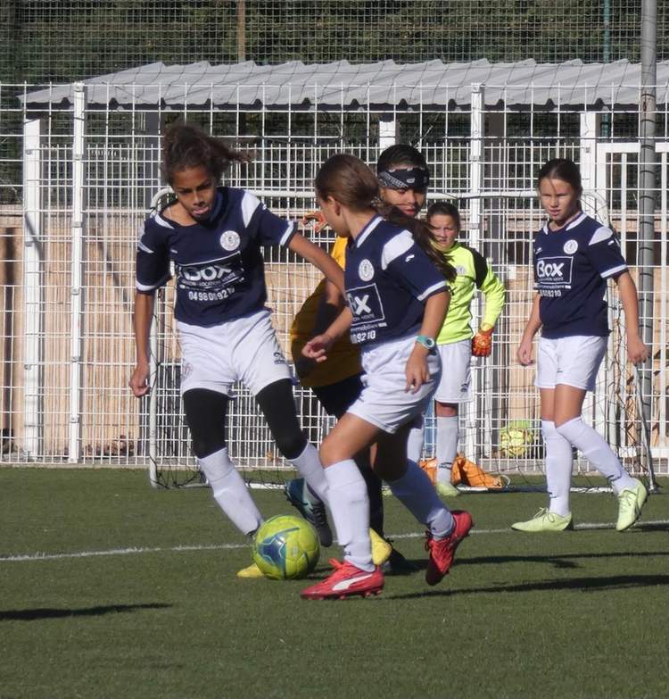 Des jeunes joueuses de football en maillot bleu et blanc sont en pleine action sur le terrain, avec un ballon au premier plan.