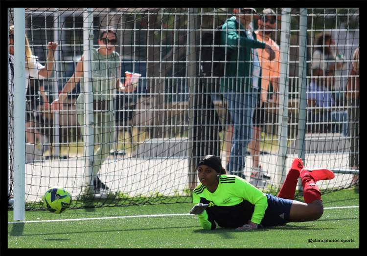 Une joueuse de football est au sol devant le but, avec un ballon à côté et des spectateurs derrière le filet.
