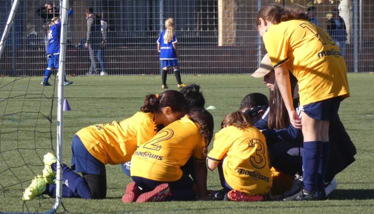 Des jeunes joueuses de football en maillot jaune et bleu sont assises ou penchées sur le terrain, avec d'autres personnes en arrière-plan.