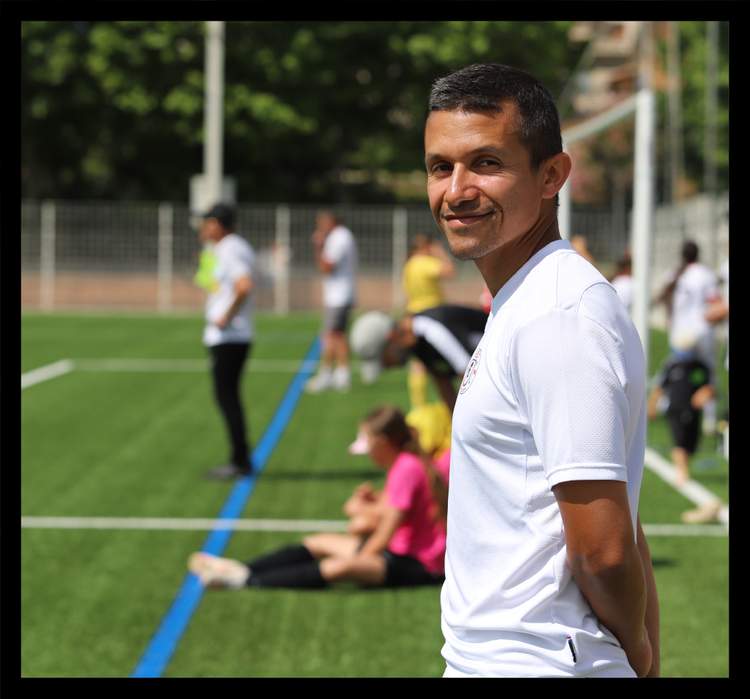Un homme souriant en maillot blanc regarde l'objectif sur un terrain de football, avec d'autres personnes floues en arrière-plan.