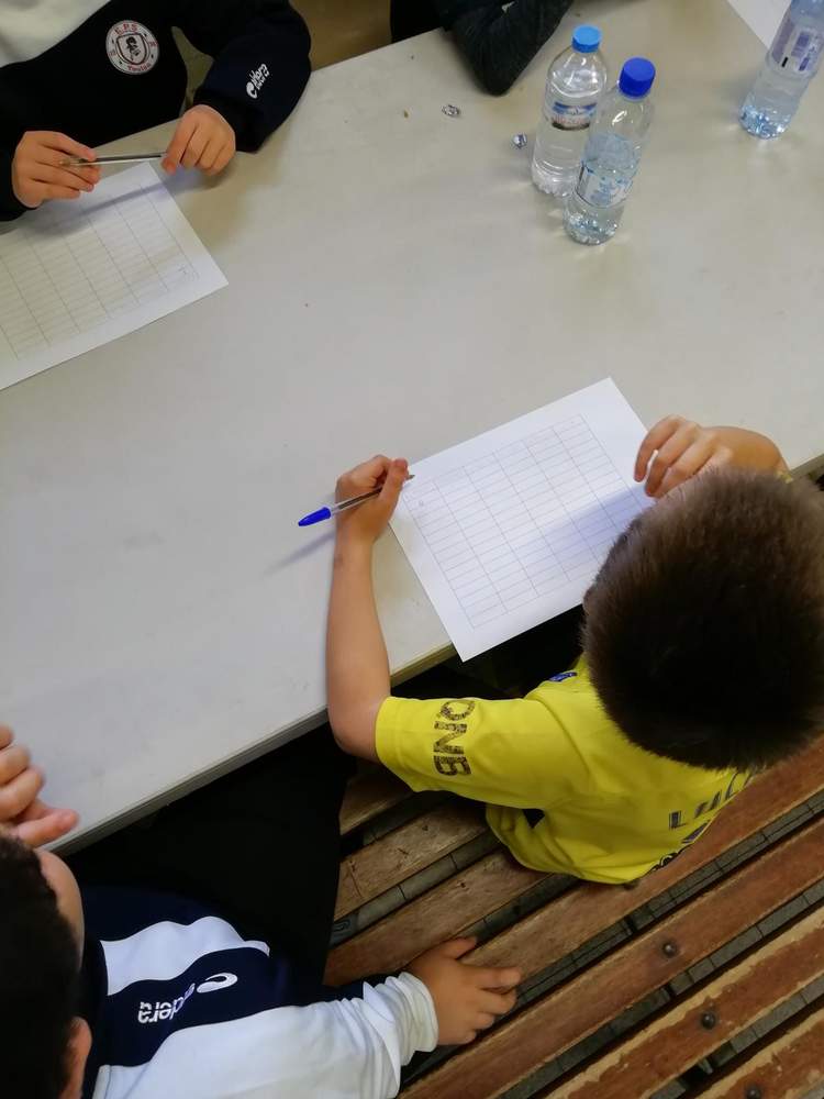Des enfants sont assis à une table et écrivent sur des feuilles quadrillées, avec des bouteilles d'eau à proximité.