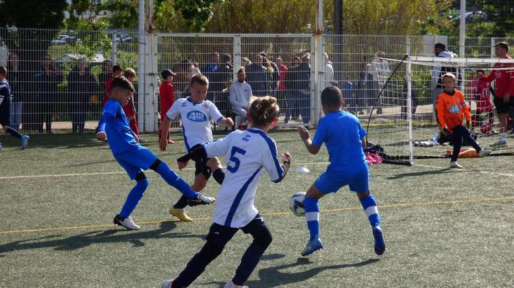 Des jeunes joueurs de football en pleine action sur un terrain synthétique, avec des spectateurs en arrière-plan.