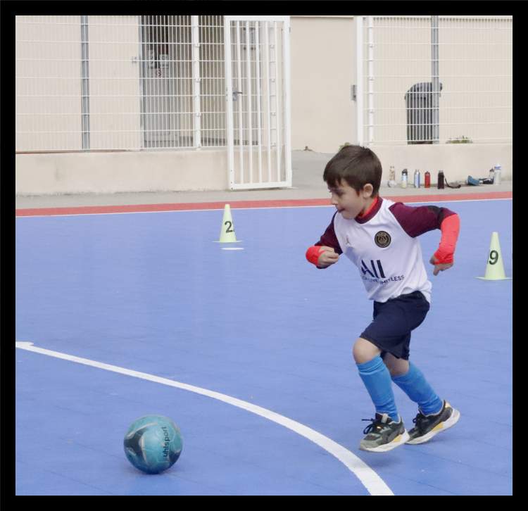 Un jeune joueur en maillot de football court sur un terrain bleu avec un ballon à ses pieds et des cônes d'entraînement en arrière-plan.