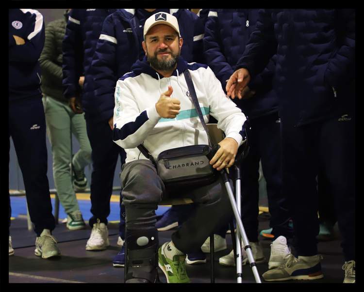 Un homme barbu souriant, assis avec une attelle à la jambe, fait un pouce levé entouré d'autres personnes.