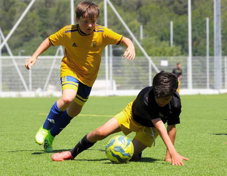 Deux jeunes joueurs de football en pleine action sur un terrain en gazon synthétique, l'un dribblant et l'autre taclant.