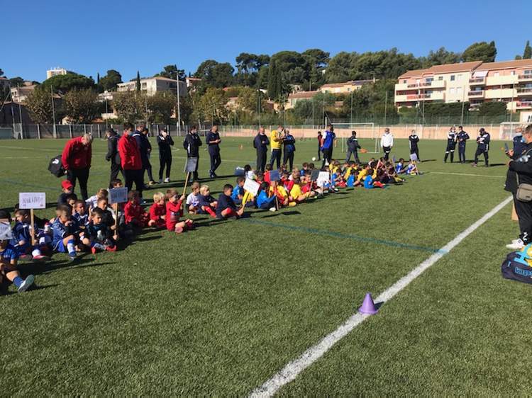 De jeunes footballeurs sont assis en rang sur le terrain, encadrés par des adultes, sous un ciel bleu.