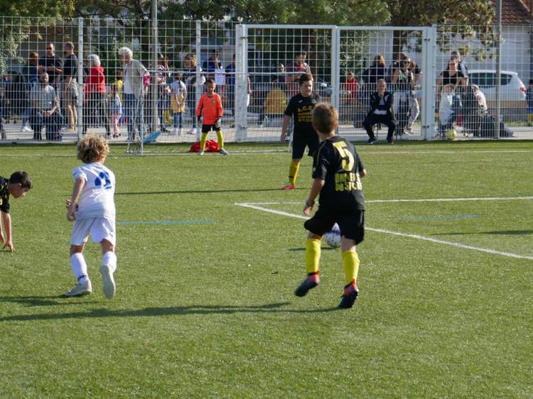 Des jeunes joueurs de football en plein match sur un terrain synthétique, avec des spectateurs derrière une clôture.