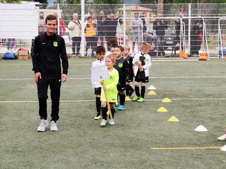 Un entraîneur et de jeunes footballeurs en tenue de sport sur un terrain, avec des spectateurs en arrière-plan.