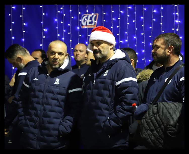 Des membres du club portent des doudounes bleues, l'un d'eux a un bonnet de Père Noël, devant un mur de lumières bleues.