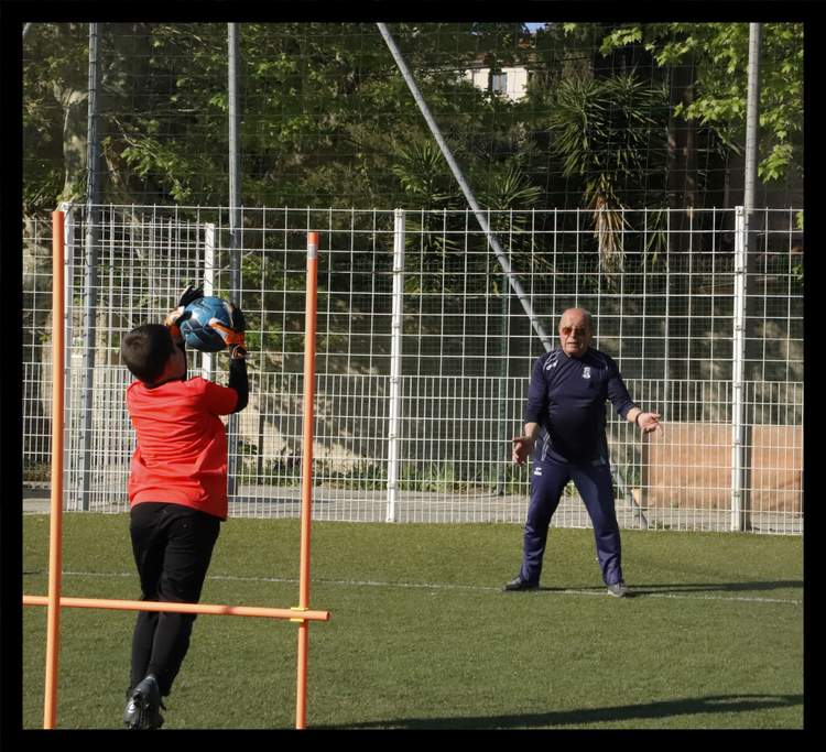 Un jeune gardien de but s'entraîne à attraper le ballon sous le regard d'un entraîneur sur un terrain de football.