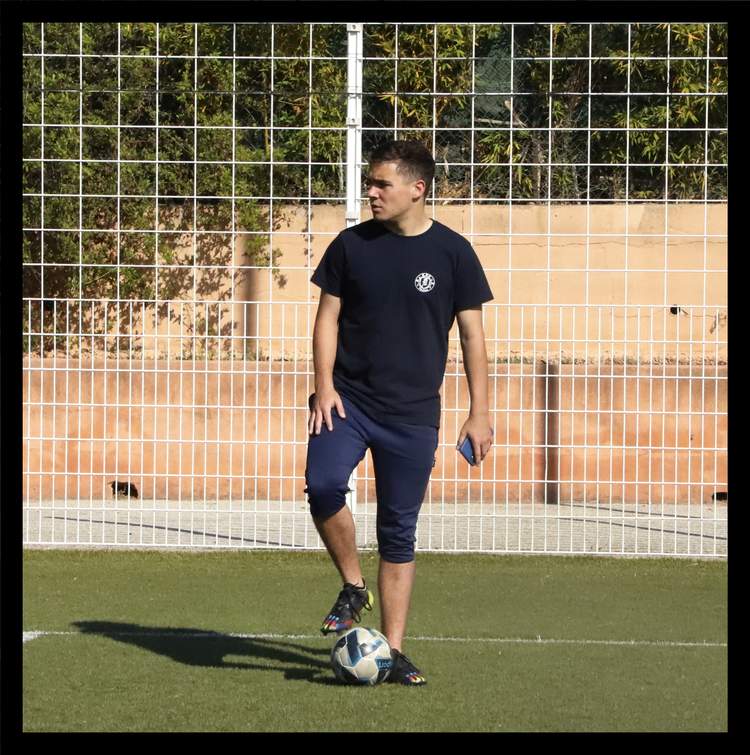 Un jeune homme en tenue de sport pose avec un ballon de football sur un terrain synthétique.