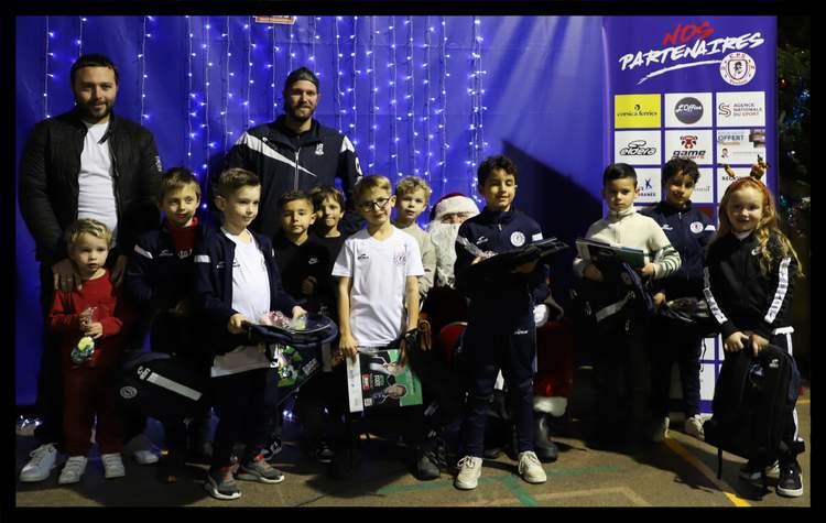 Des enfants et des adultes posent avec le Père Noël devant un mur décoré de lumières et une bannière de partenaires.