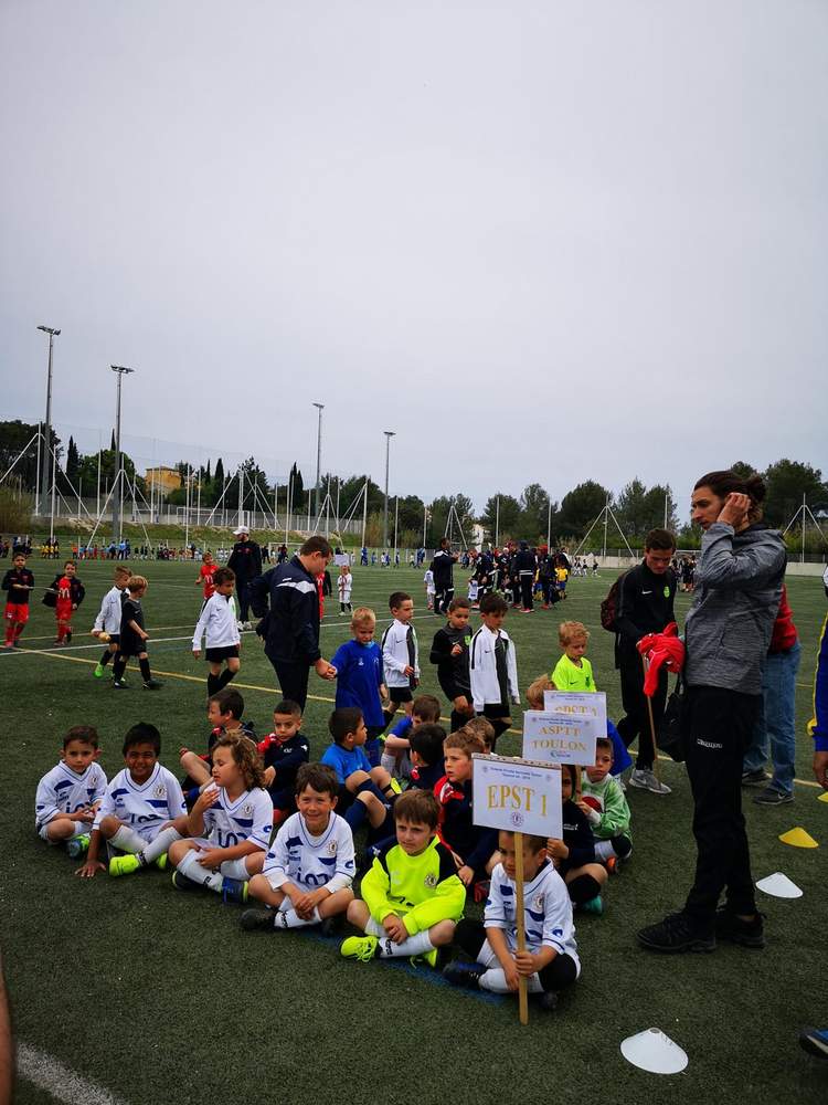 De jeunes footballeurs sont assis sur le terrain, attendant le début d'un match ou d'un événement, avec des adultes et d'autres enfants en arrière-plan.