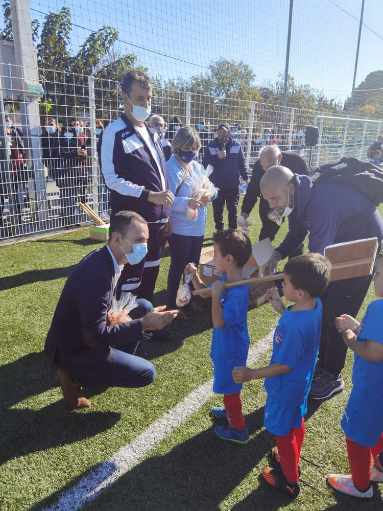 Des adultes masqués et des enfants en tenue de football participent à un événement sur un terrain de sport en gazon synthétique.