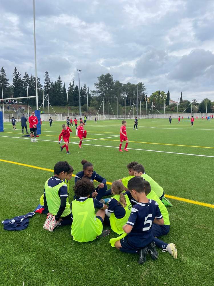 Des jeunes footballeurs en maillots de différentes couleurs sont assis ou courent sur un terrain de football synthétique sous un ciel nuageux.