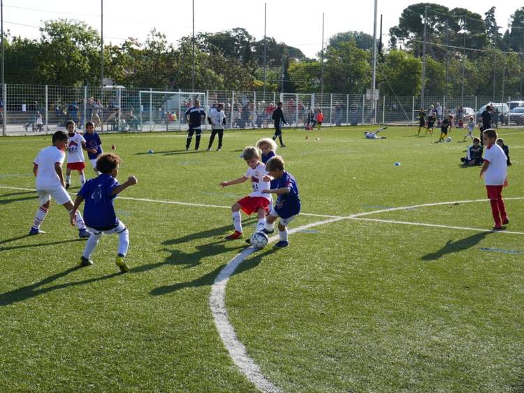 Des jeunes joueurs de football en pleine action sur un terrain ensoleillé, avec d'autres enfants et adultes en arrière-plan.