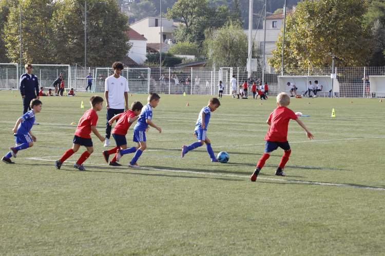 Des jeunes joueurs de football en maillots rouges et bleus s'entraînent sur un terrain ensoleillé, sous la supervision d'adultes.