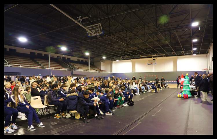 Une foule de personnes, dont de nombreux jeunes en survêtement, assiste à un événement dans un gymnase décoré pour Noël.