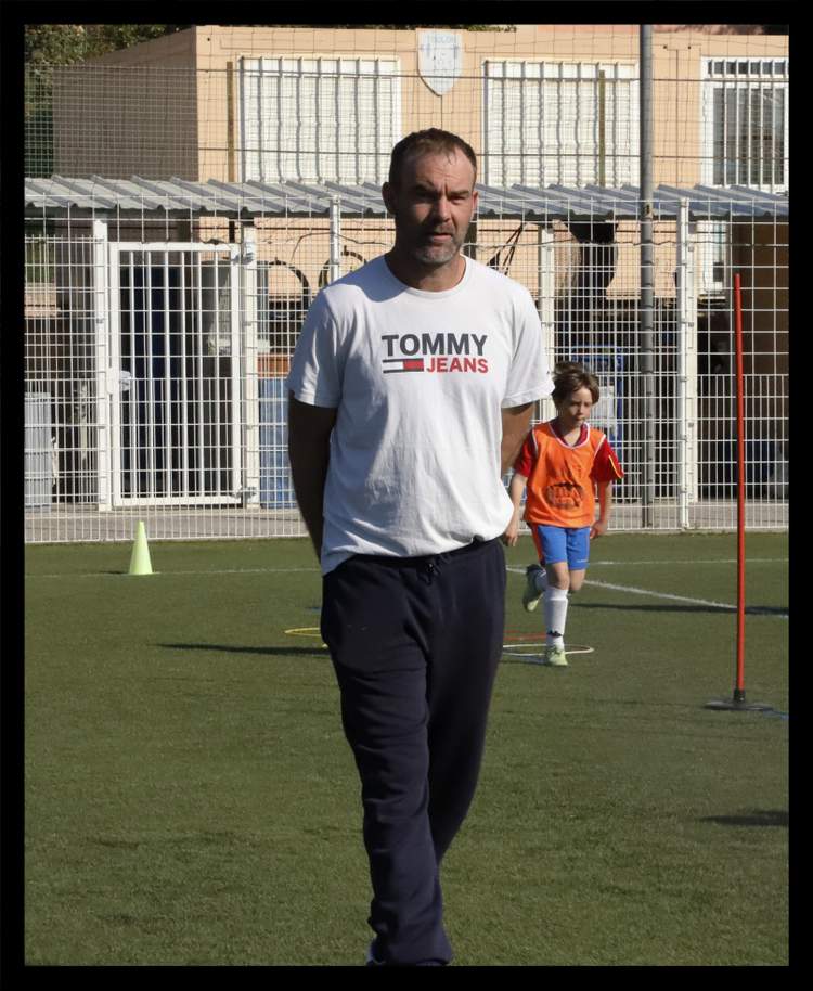 Un entraîneur adulte en t-shirt blanc et un jeune joueur en orange sur un terrain de football.